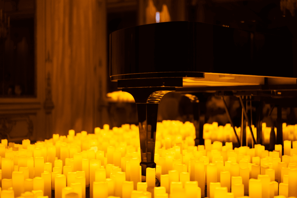Concert Candlelight au piano, beaucoup de bougies au premier plan, gros plan sur le pied du piano avec un fond noir et le lumière des flammes qui se reflète sous le piano.